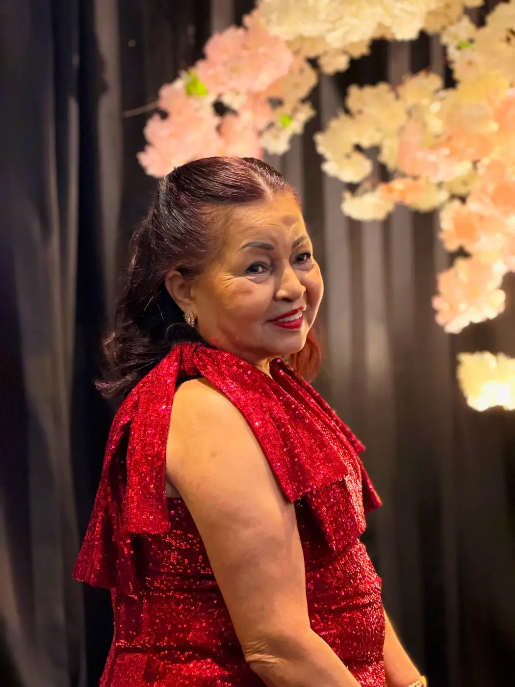 Aniversariante de vestido vermelho de paetês sorrindo diante das flores